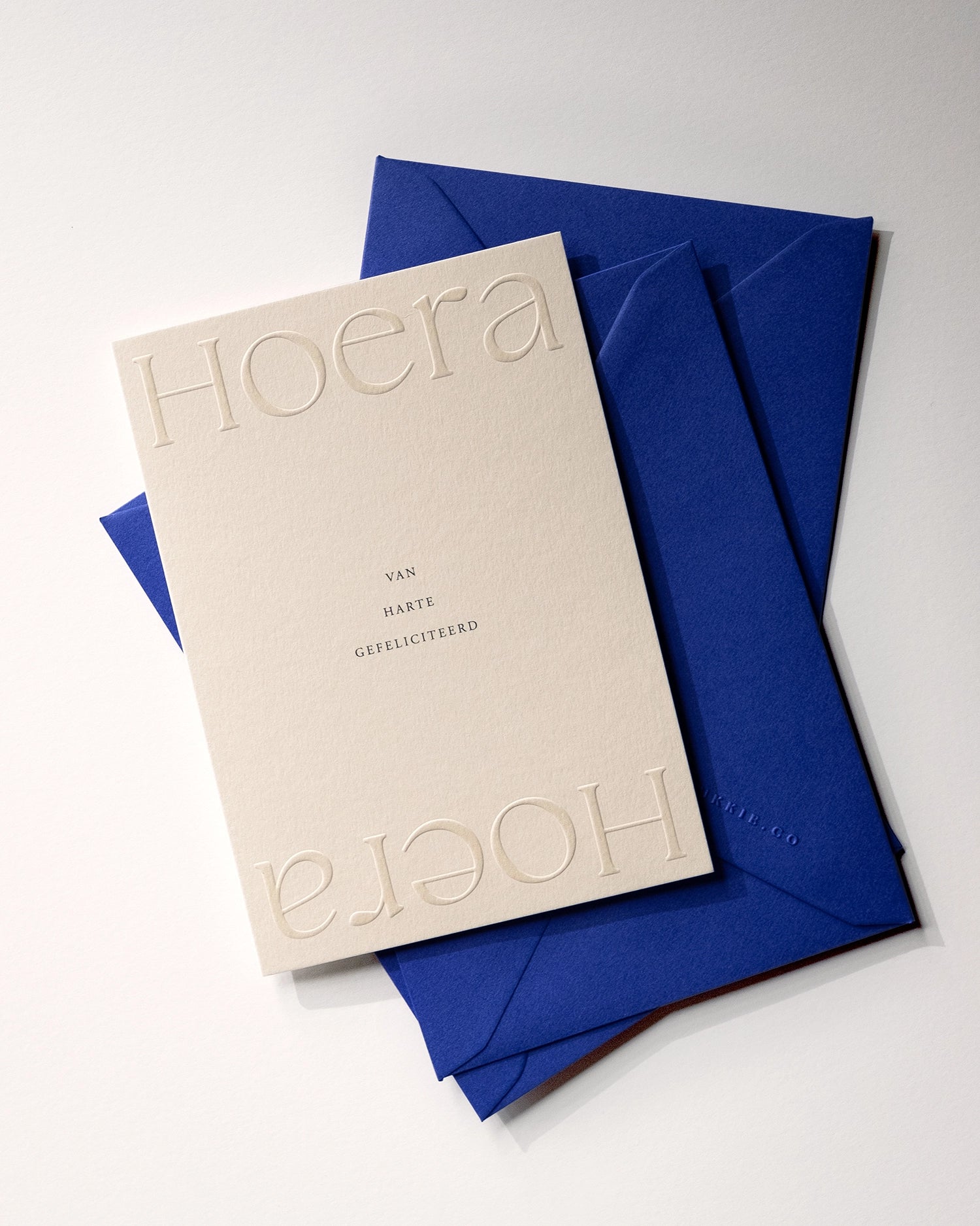 Flat lay of minimal luxury embossed birthday card “hoera van harte gefeliciteerd” on stack of playful royal blue envelopes