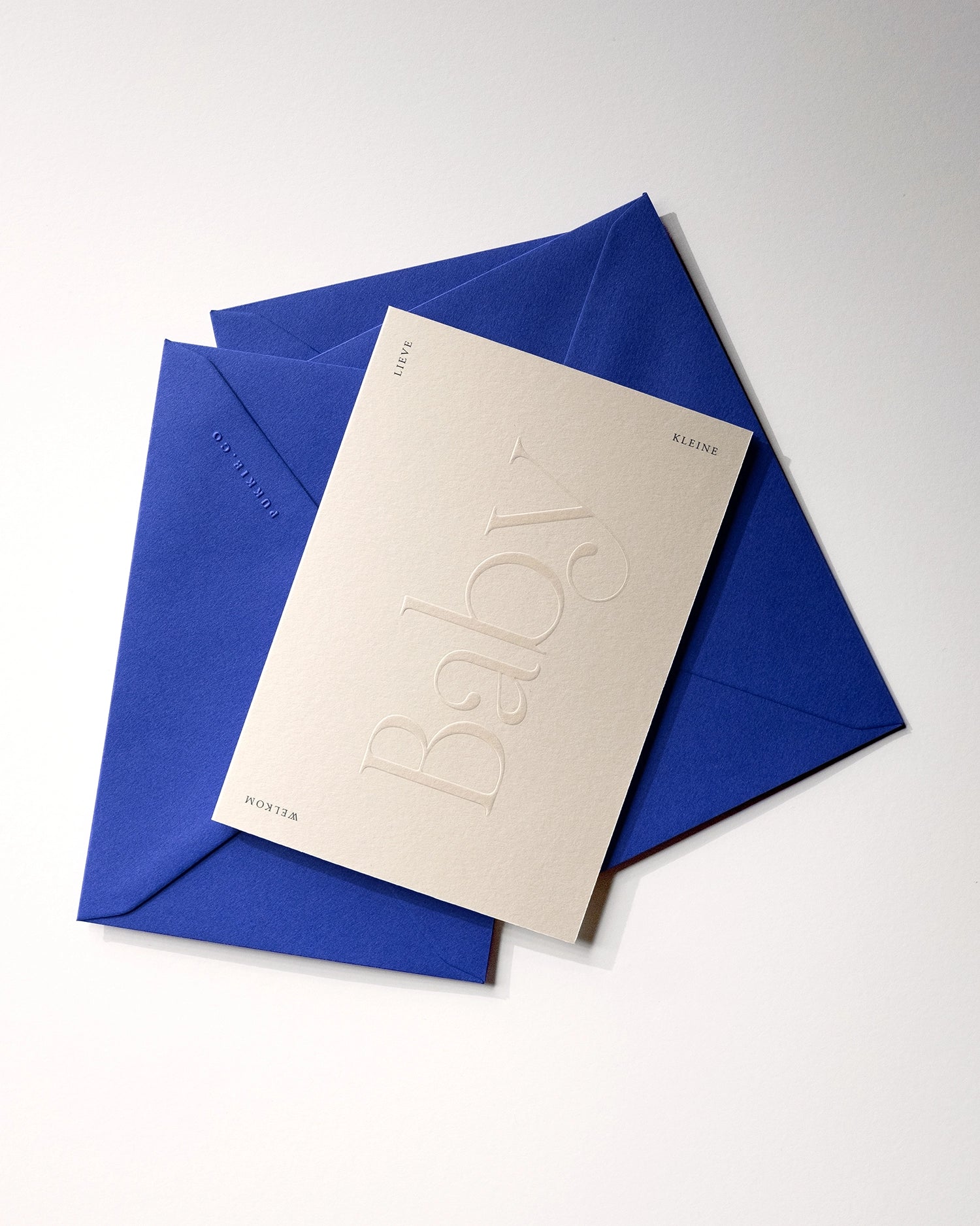 Flat lay of minimal luxury embossed new baby card “welkom lieve kleine baby” on stack of playful royal blue envelopes