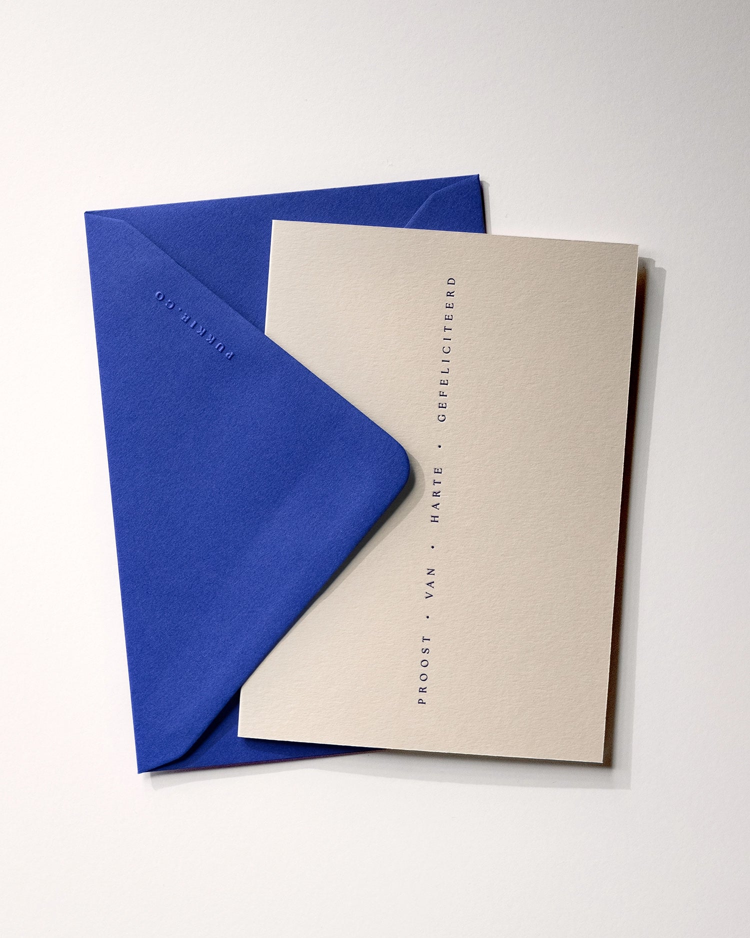 Flat lay of minimal luxury letterpress celebration card “proost van harte gefeliciteerd” with envelope folded over card on Colorplan Mist paper